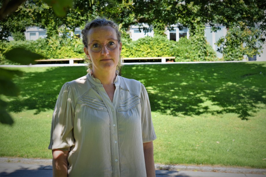 Kvinne med briller og beige bluse står ute i grønne omgivelser.