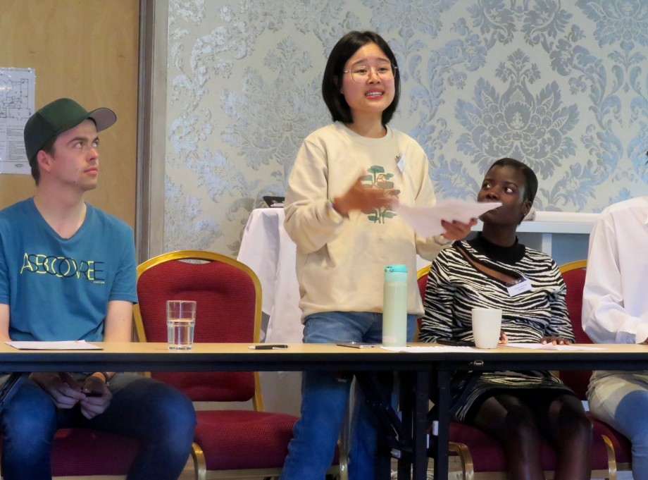 Tre studenter i rollespill bak et skrivebord. En ung mann sittende til venstre med caps og blå t-skjorte, en ung kvinne stående i midten, snakkende med et ark i hånden og en ung kvinne sittende til høyre. Foto.