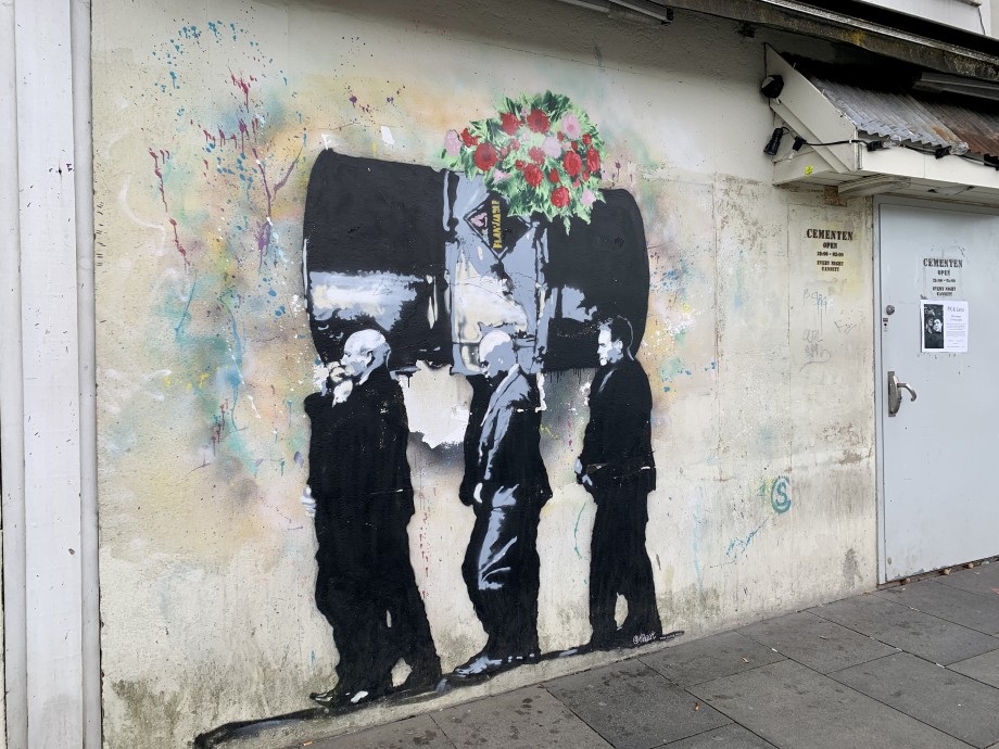 Graffit of six men carrying an oil drum on their shoulders as if bearing a coffin at a funeral. A wreath of flowers has been placed on the oil drum