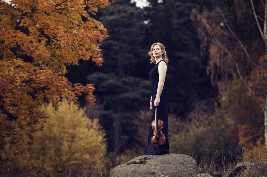 Fotografi av kvinne som holder en fiolin. Har på seg en lang sort kjole, og står på en stein i høstlig landskap i skogen.