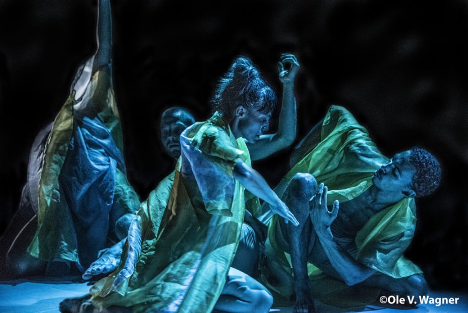 Fotografi av uttrykksfull samtidsdans. 4 personer i kapper som sitter på gulvet i ulike dansepositurer på en mørk scene med blått lys.