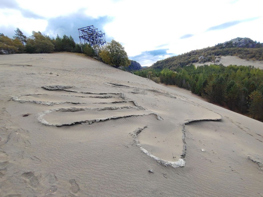 Bilde av et mønster i en sandbak. Et stativ, skog og himmel i bakgrunnen. Foto.