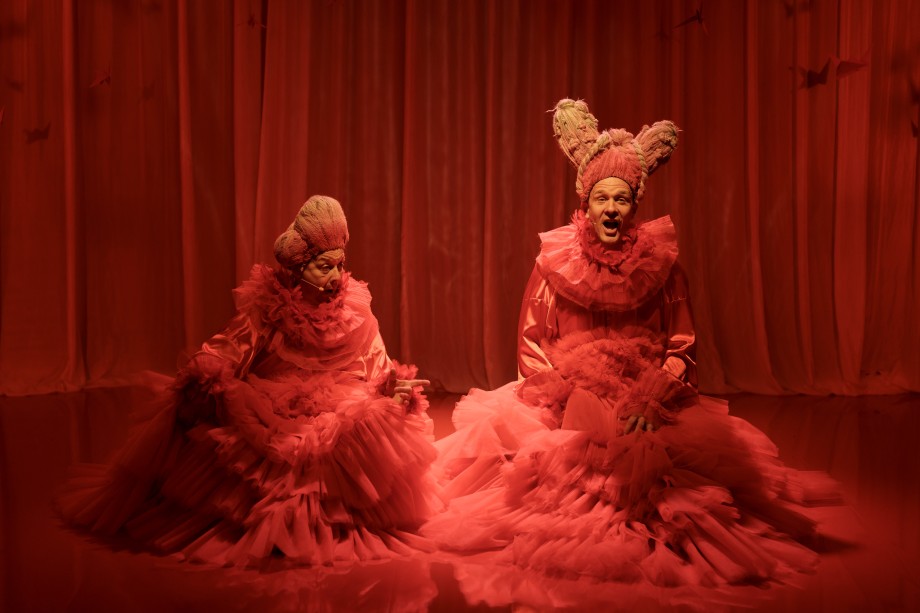 To menn som sitter på gulvet på en scene og synger i overdådige barokke kjoler og parykk. Foto.
