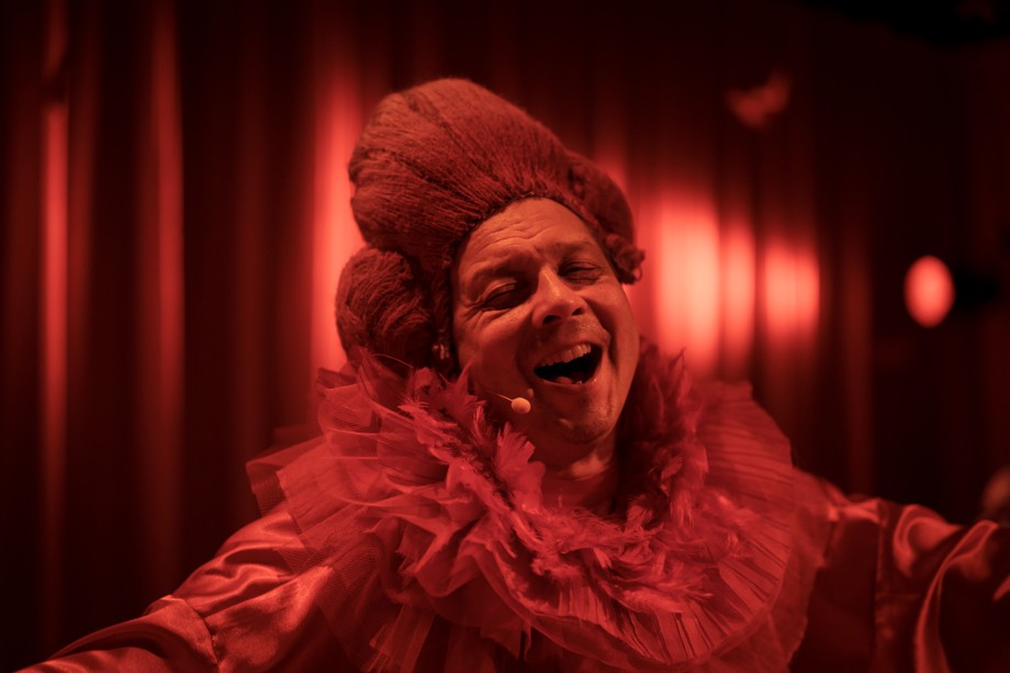 Smilende mann som synger med øynene lukket i overdådig rød parykk og kjole. Foto.