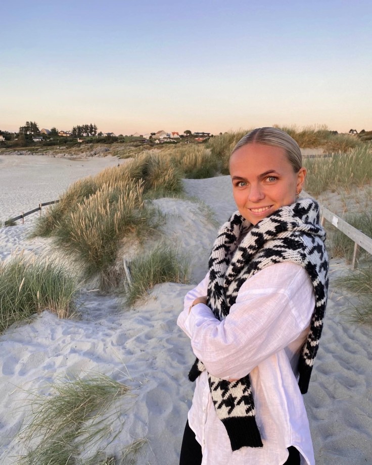 ung kvinne med lyst hår står på en sandstrand i solnedgang. Hun har på seg en hvit skjorte og en genser over skuldrene og smiler til kamera.