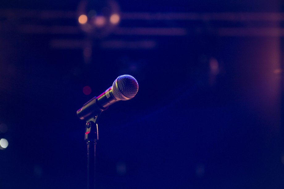 Nærbilde av en mikrofon på et stativ i et lite belyst rom med scenelys i bakgrunnen.