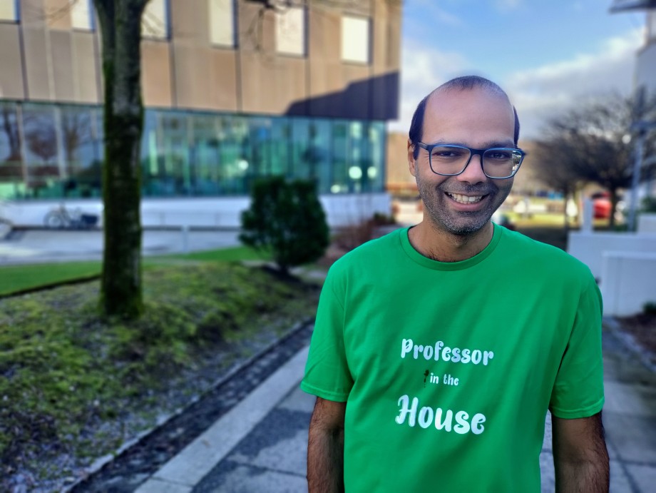Foto av en smilende mann med briller i ført en grønn t-skjorte hvor det står "Professor in the House" står utendørs med blå himmel og en sort bygning i bakgrunnen.