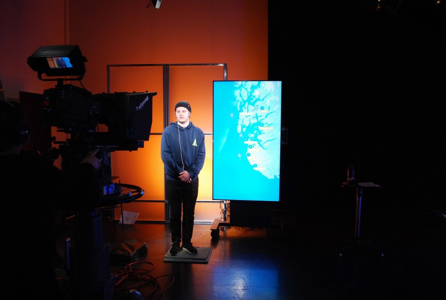 Ung mann står foran et TV-kamera med en skjerm med et kart ved siden av seg og oransje vegg bak seg.
