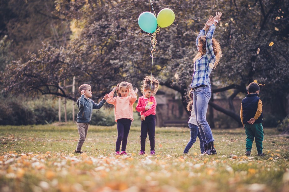 Fem barn og en voksen utendørs. Ett av barna holder to ballonger. Den voksne hopper høyt i været.