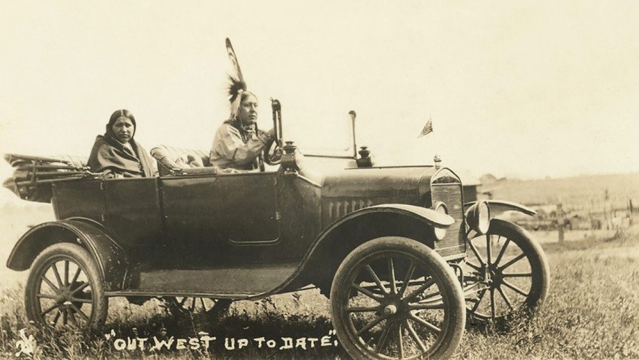 A black-and-white photograph of two members of the Osage Nation, one wearing a feathered headdress and the other in a shawl, sitting in a Ford Model T. A hand-written caption beneath reads: “Out West and Up to Date.”