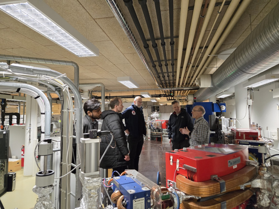 People standing in a ring among industrial equipment for a heating facility. One of them is explaining the facility to the rest