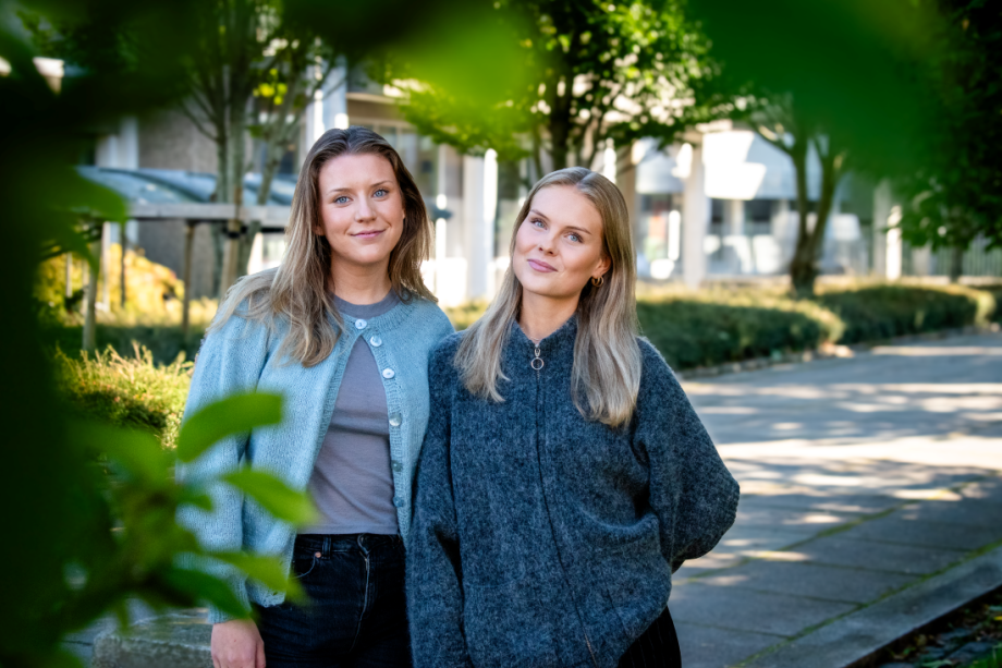 To unge kvinner står ute omkranset av grønne blader. De ser inn i kamera og smiler.