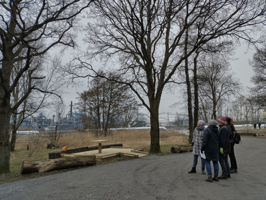 A group of four people stand on a path in a park looking through the trees towards an oil refinery in the background