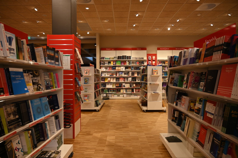 Fotografi tatt fra innsiden av bokbutikken Akedemia - viser hyller fylle av bøker på høyre og venstre siden. Og i enden av bildet. To store røde søyler til høyre og venstre i bildet.