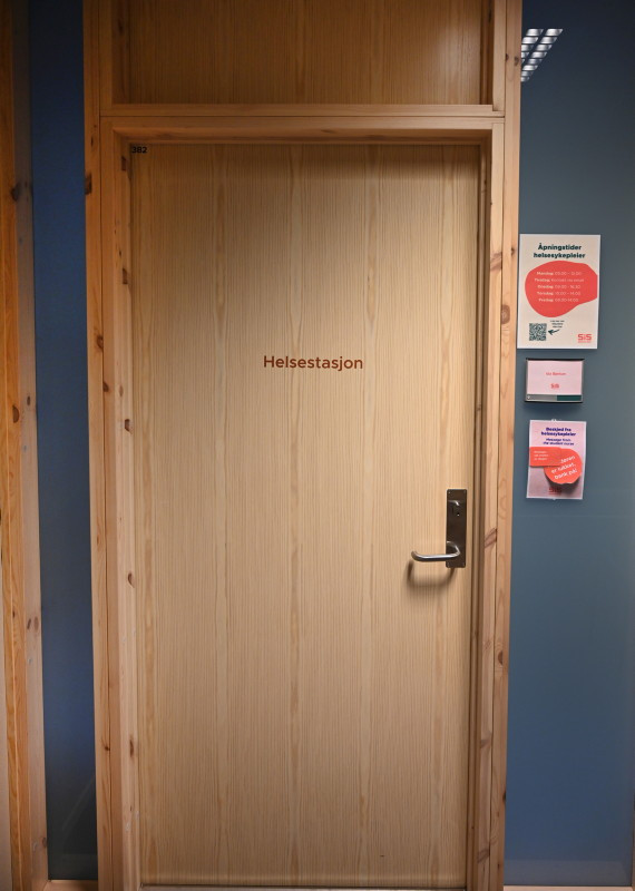 Fotografi av døren til Helsestasjonen i tredje etasje på SiS Studenthus - døren er beige/lysebrun det står "helsestasjon" på øverste halvdel av døren i rød skrift - ved siden av skriften på beggen henger det noen fargerike lapper og plakater med informasjon.