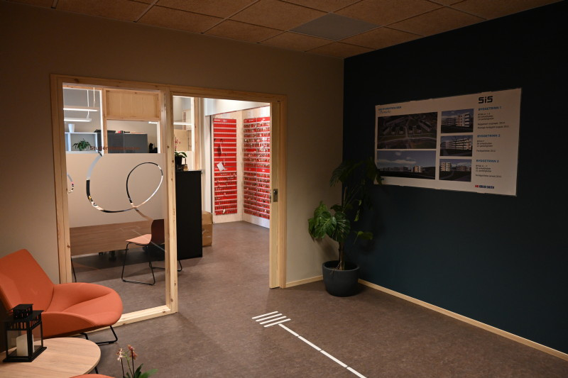 Fotografi av Studentboligkontoret tatt fra heisen. En stol og bord til venstre i fotografiet. kontoret rett frem med en vegg av nøkler og en inngang. til høyre i bildet en vegg med en plakat om byggeprosjekter.