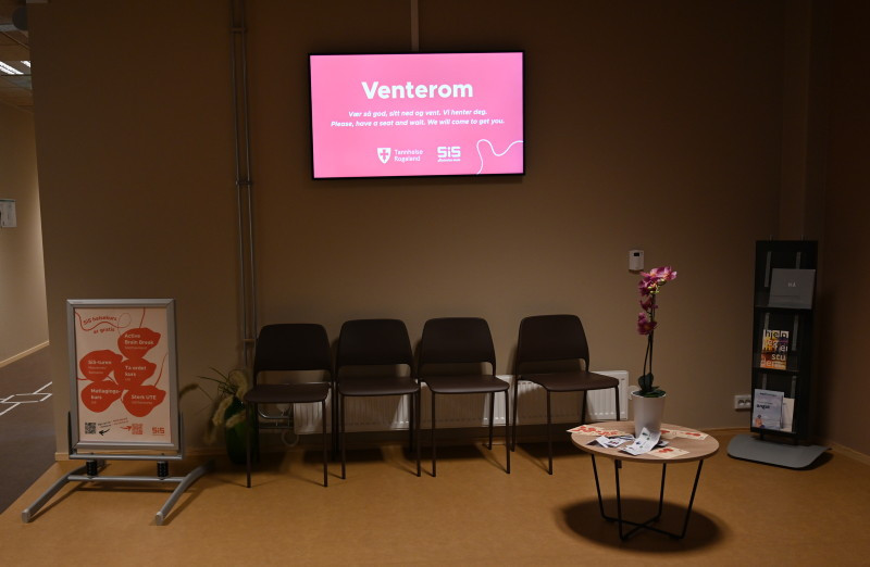 Fotografi av venterommet til Rogaland tannhelseklinikk på Ullandhaug, SiS Studenthus. Fire svarte stoler i bakgrunnen av bildet. Et lite bord ved siden av til venstre, og til høyre et stående skilt med informasjon. Ovenfor stilene på veggen er det en tv-skjerm med informasjon. Helt til høyre i rommet en stående metall magasinholder.