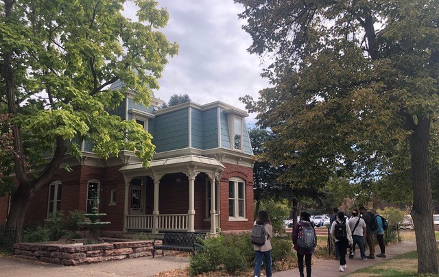 A colonial style house with red brick walls and blue-grey tiles sits in the centre of the picture with trees flanking it. Students with backpacks are walking past it away from the camera