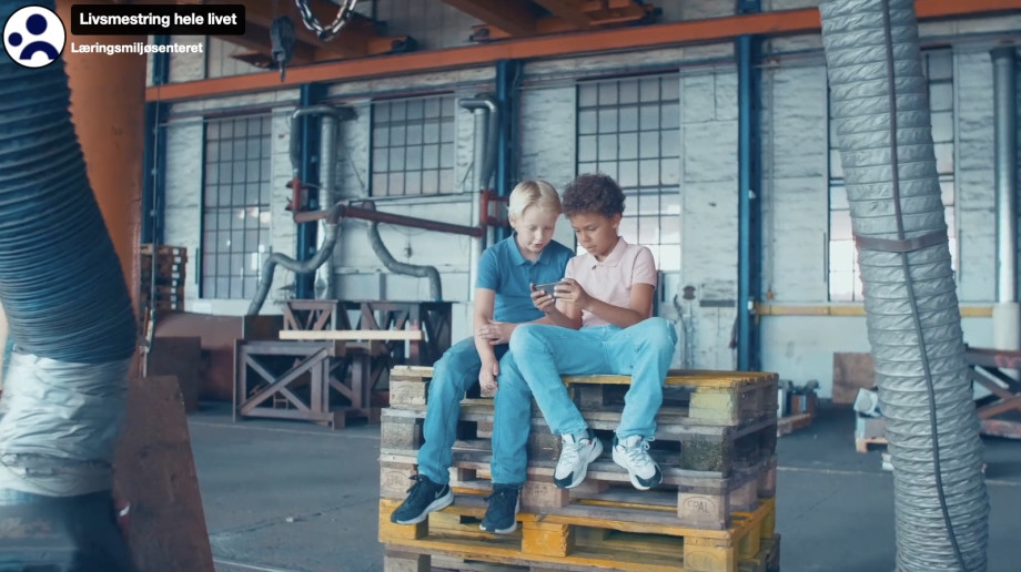 Skjermdump fra filmen Livsmestring hele livet: To gutter sitter på en stabel paller og ser på en mobil sammen.