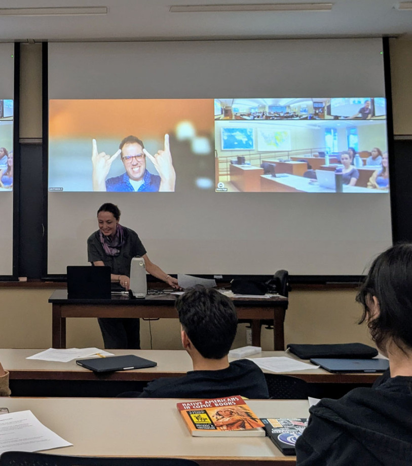 A screen at the back of a classroom displays a male lecturer presenting digitally to a classroom of students. We see the screen from the perspective of one of the students. The moderator stands front left of the screen and is smiling at the laptop in front of her