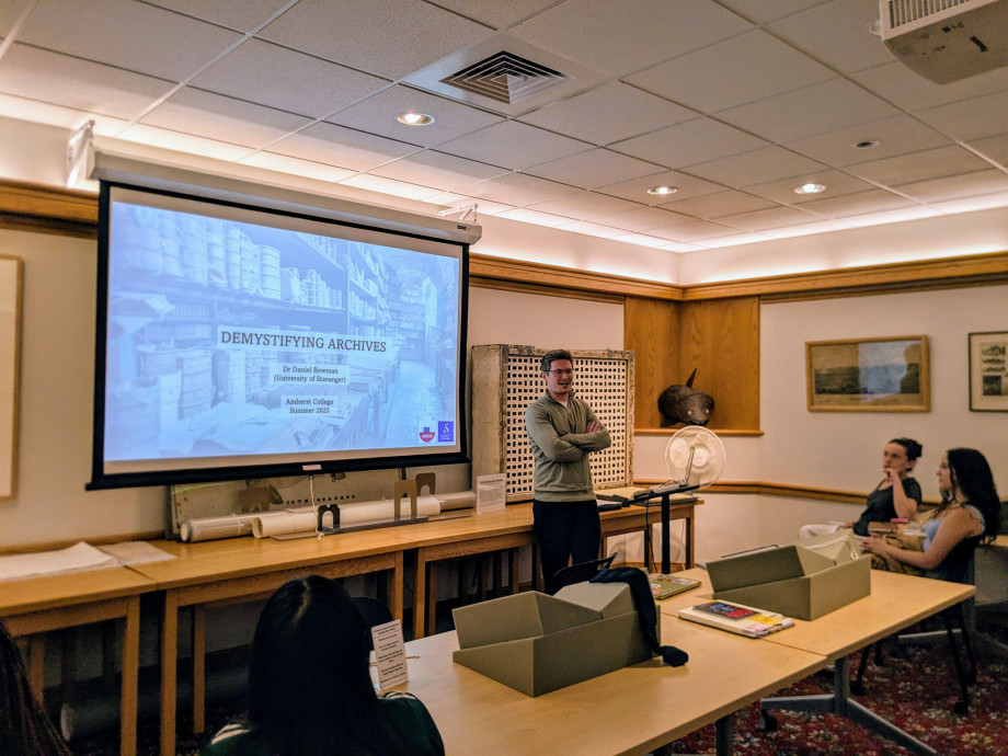 A classroom. The opening slide of a presentation can be seen on the screen in the centre of the picture. The lecturer stands in front of it while seated attendees are looking at him