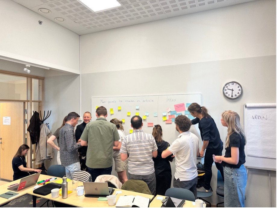 A group of people are gathered around a whiteboard with post-it notes on it. They are in animated discussion