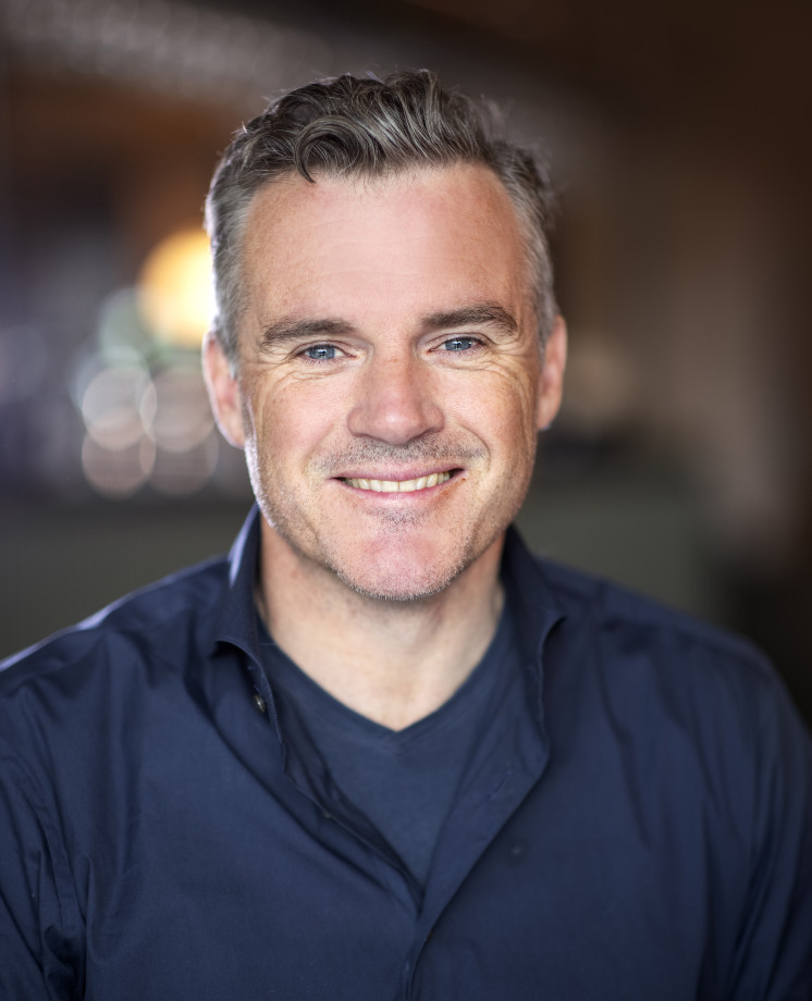 Mann i skjorte smiler mot kamera. Uskarp bakgrunn.