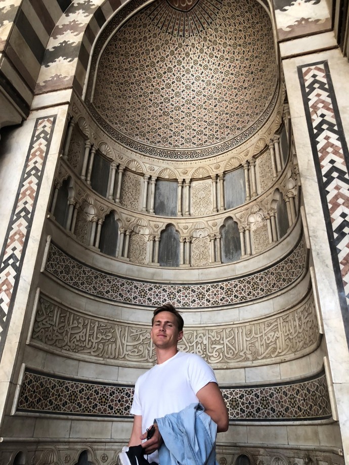 Johannes midtsentrert, bildet tatt nedenfra. Han er iført en hvit t-skjorte med en lys, tynn og blå dongeriskjorte over armen. Bak han ser man deler av innsiden på Ibn Tulun-mosekeen. Han står inni et slags halvt sylinder med fargerike fliser og skriftspråk.