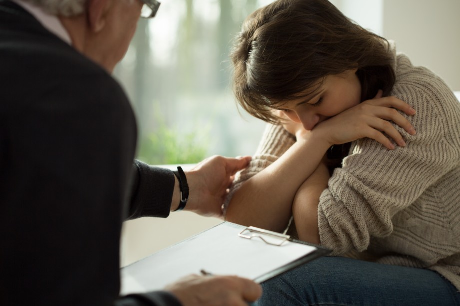 En voksen trøster en ung jente som sitter med bøyd hode og armene rundt seg.