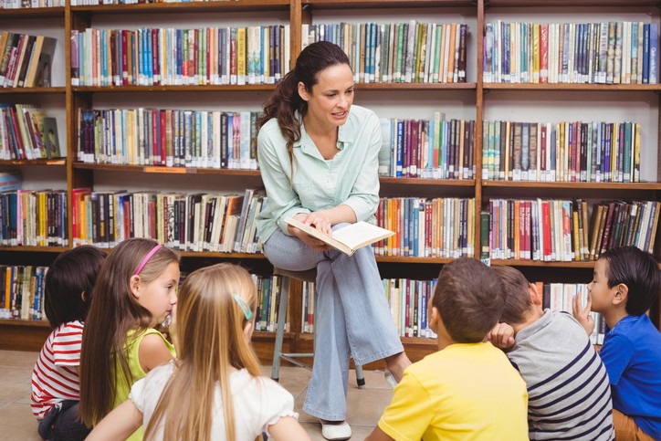 Lærer  eller bibliotekar leser for flere barn. Foran en stor bokhylle.