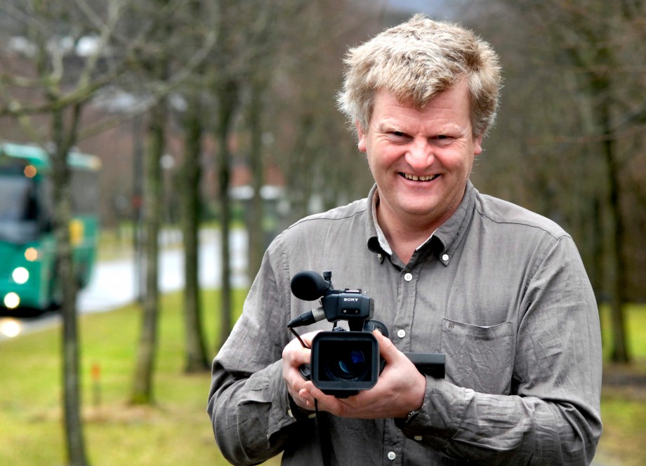 Hans Eirik Voktor smiler og holder et filmkamera. Han står ute med trær og UiS i bakgrunnen.