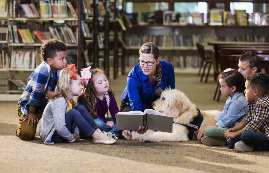 En gjeng med barn og en hund sitter på gulvet og lytter til en lærer som leser.