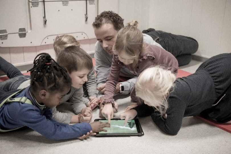 Barn og barnehagelærer ser på nettbrett på gulvet.