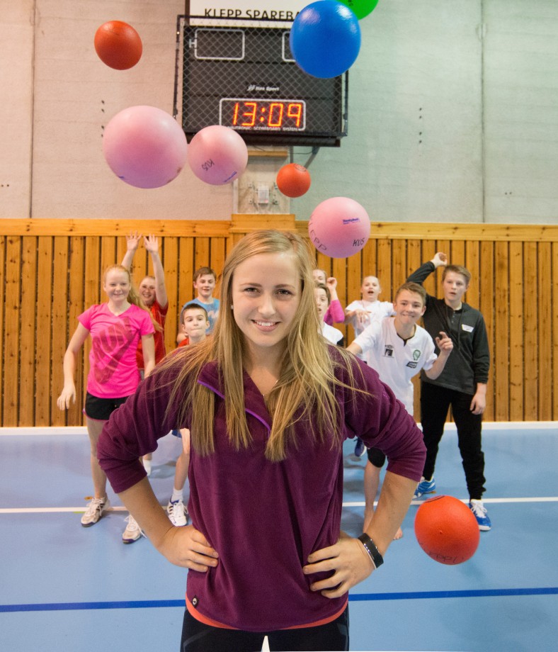 ung kvinne står i forgrunnen, ser i kamera og smiler, er kledd i sportsklær. Bak henne holder barn på med ballspill. 