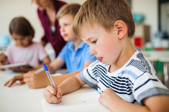 Skolebarn skriver og tegner i klasserom, lærer i bakgrunnen