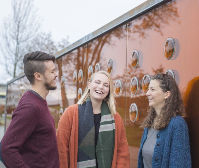 Glade studenter foran bronseveggen på Ullandhaug campus,
