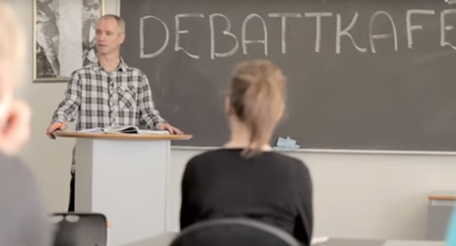 Lærer står foran tavle der det står debattkafe. Snakker til klassen. 