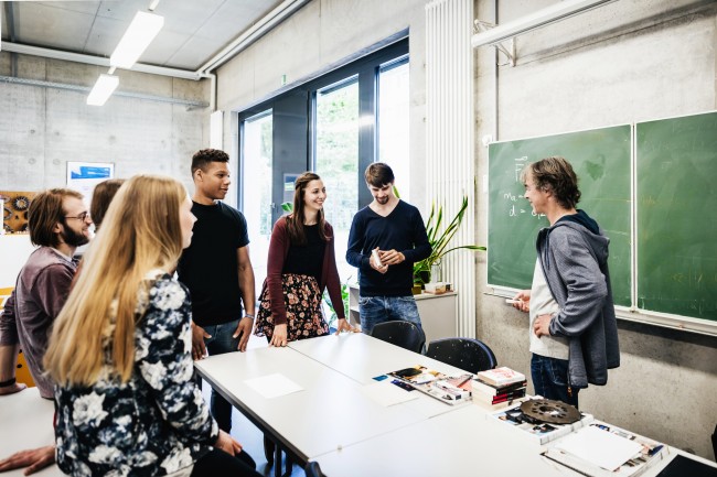 En lærer og en gruppe elever står i et klasserom og snakker sammen.