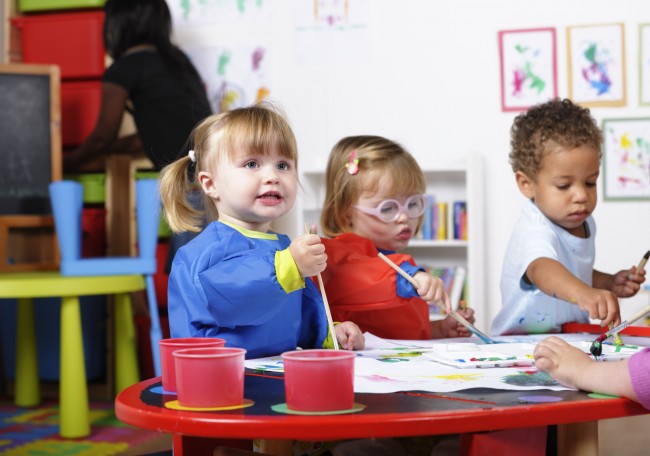 tre små barn i fargesterke klær sitter rundt et bord og maler 