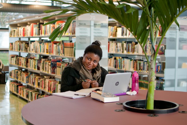 Dame sitter ved rundt bord med palmeplante og studerer på en PC