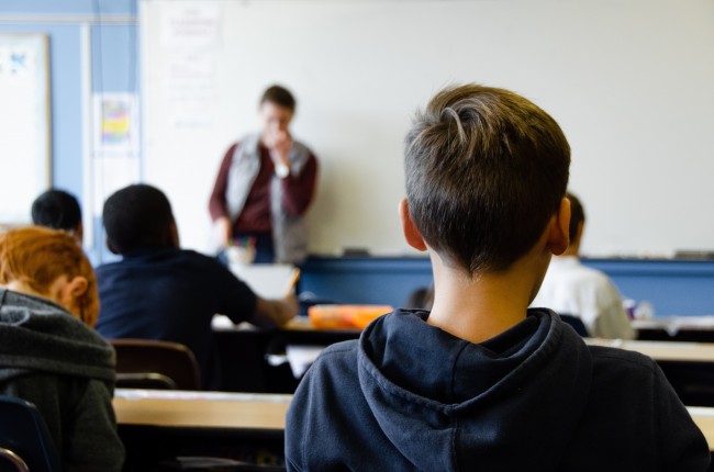 Bakhodet til en gutt. Lenger framme ser vi flere elever ved skolepulten og en lærer foran en hvit tavle.