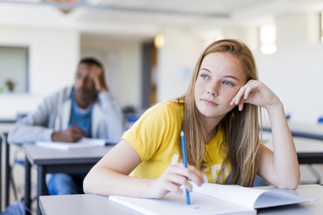 Jente sitter ved pulten sin i et klasserom med en skrivebok foran seg og pennen i hånden. Det ser ut som om hun tenker på noe