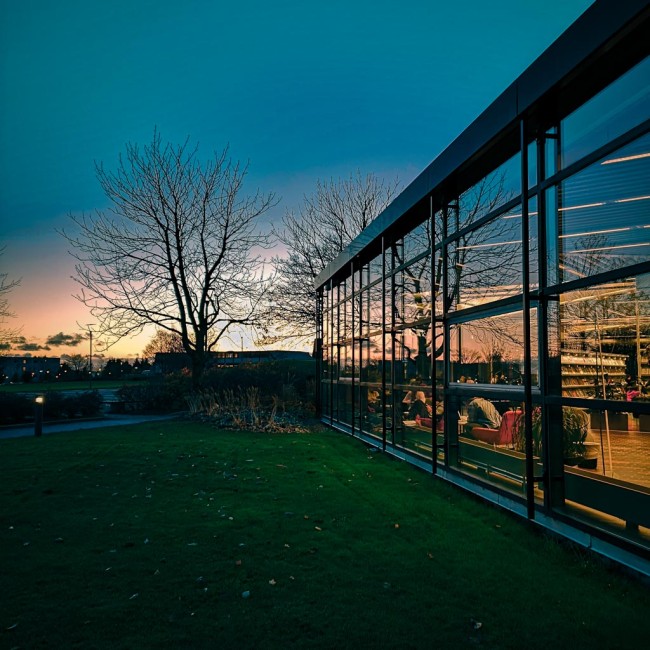 Bygning med store glassvindu som reflekterer solnedgangen i horisonten og mennesker på innsiden som leser bøker