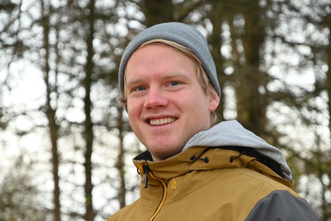 En ung mann med grå lue og gul jgakke med hette står i skogen og smiler til kamera