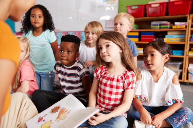 En gruppe på sju barn sitter og hører på en lærer som leser i en bok