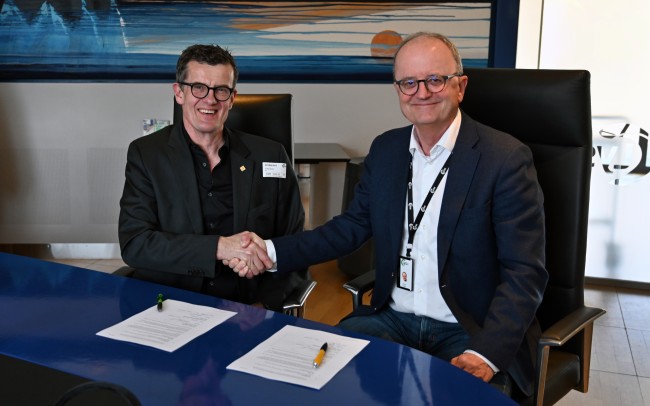 Klaus Mohn og Eimund Nygaard tar hverandre i hånden ved et styrebord med to signerte ark foran seg.