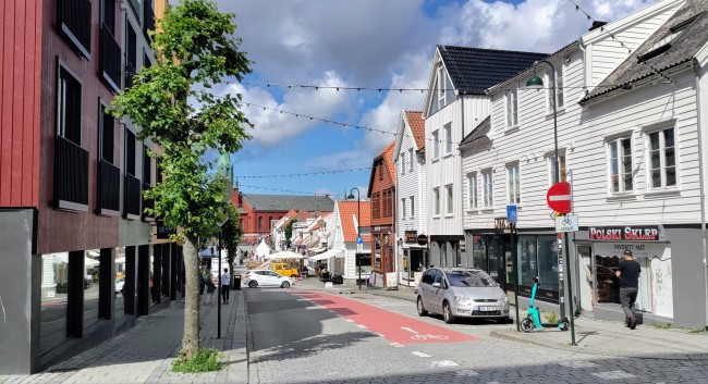 Pedersgata i Stavanger med hvite og oransje hus på høyre side og en rød bygning på venstre side. Et grønt tre står på venstre side, og en grå bil er parkert på høyre side. En mann går på høyre fortau. Foto.