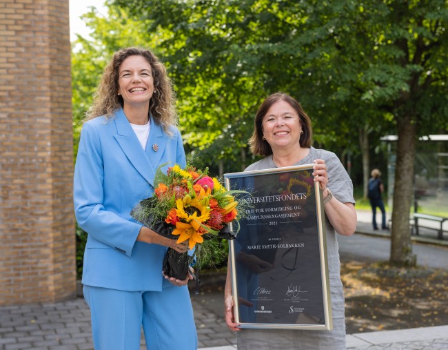 Marie Smith-Solbakken (t.h.) med diplom og Cecilie Claviez med blomsterbukett foran stort grønt tre