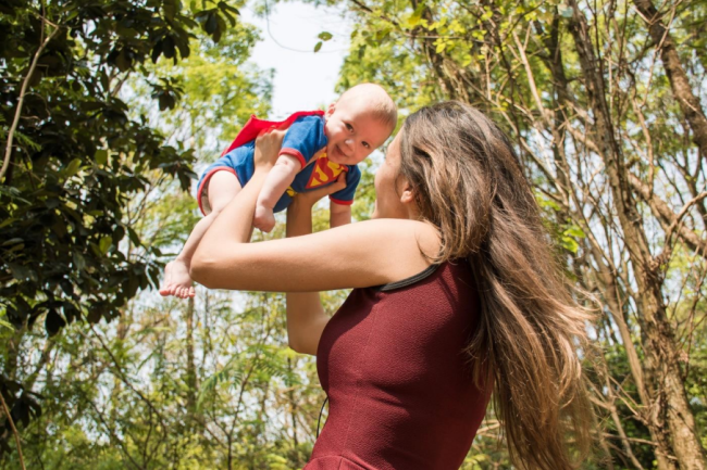 Kvinne med ryggen til holder opp baby som smiler iført supermann-body