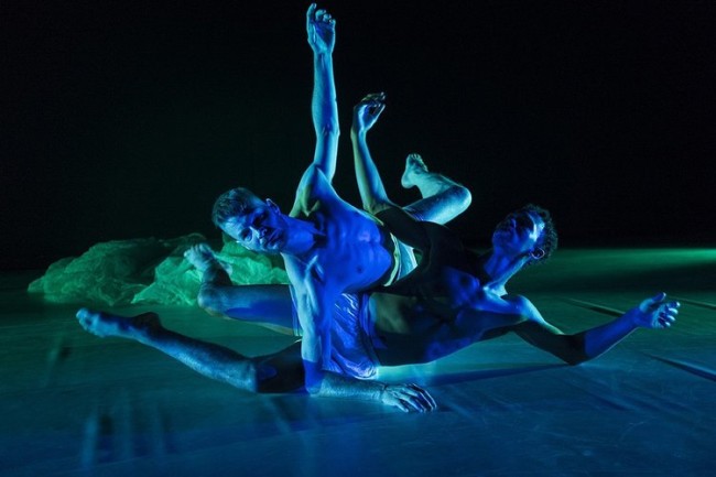 Fotografi av to dansere på en mørk scene som ligger på gulvet, hvor han ene ligger oppå sidelengs på han andre.